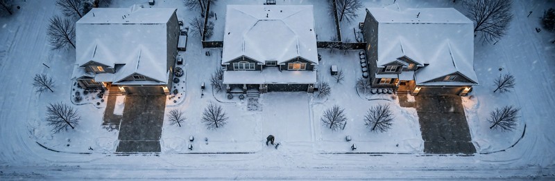 Overhead view of a man shoveling a single straight path through deep snow to his open garage, neighbors' driveways and sidewalks cleared, street snow-covered.